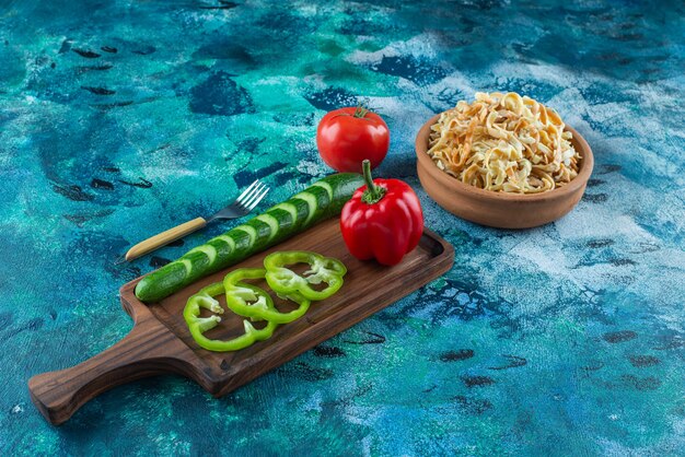 Variously vegetables on a board next to fork an a bowl of noodle on the blue table. 