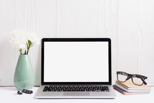 Vase; paperclip; laptop; notebooks and eyeglasses against painted white wall