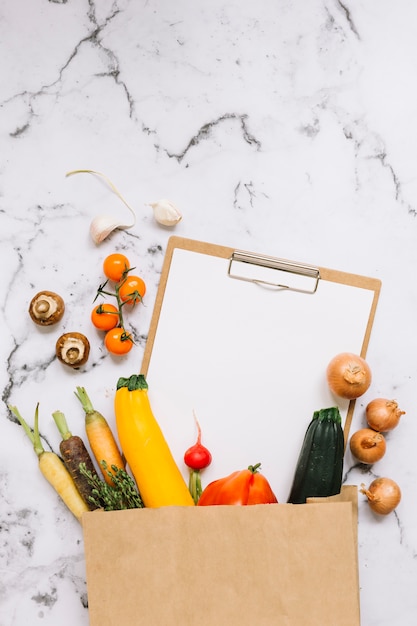 Vegetables and paper on clipboard falling from brown paper bag on marble background