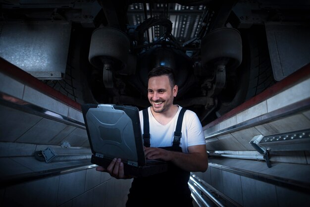 Vehicle mechanic with diagnostic tool laptop working under the truck in workshop