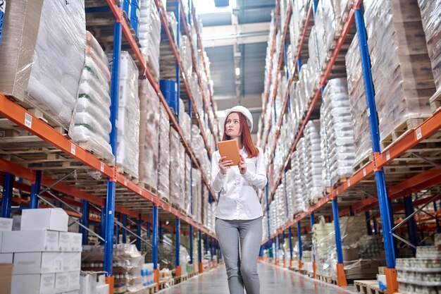 Verification. Young long-haired woman in white protective helmet with tablet in hands looking attentively at shelves with goods in warehouse walking in aisle