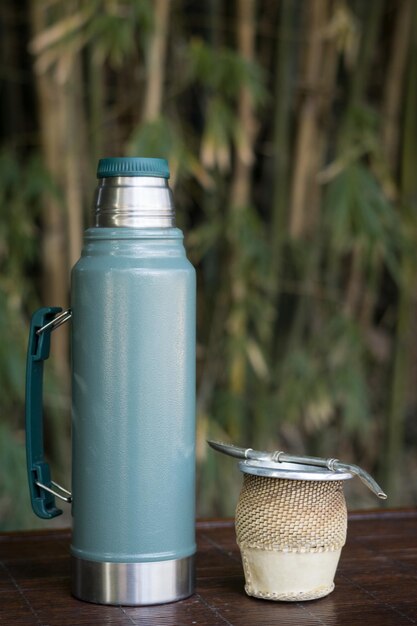 Vertical closeup of a cup of yerba mate infusion with a thermos.
