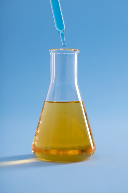 Vertical closeup of dropper with blue liquid over Erlenmeyer flask yellow liquid on blue surface