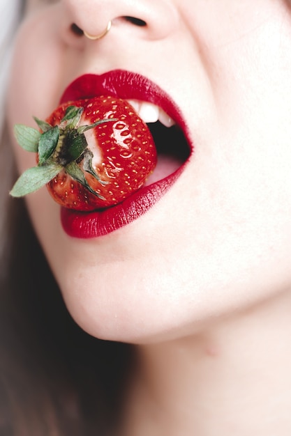 Vertical closeup shot of a young female with red lipstick biting a strawberry