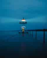 Free photo vertical distant shot of a person holding an umbrella walking on a footbridge near a lighthouse