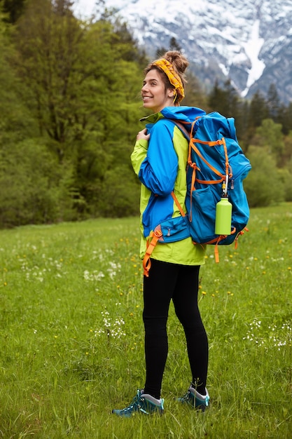 Vertical full length shot of pleased active female tourist dressed in active wear, wanders on green meadow