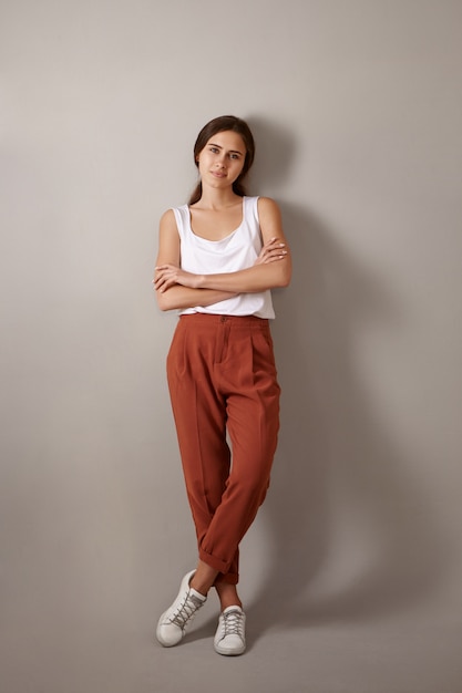 Vertical full length trendy looking fashionable young European woman dressed in white top, brown trousers and sneakers posing isolated, crossing arms confidently, enjoying leisure time