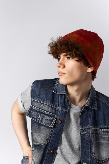 Vertical image of attractive trendy young Caucasian male wearing red hat and stylish denim vest over gray t-shirt looking away with pensive facial expression