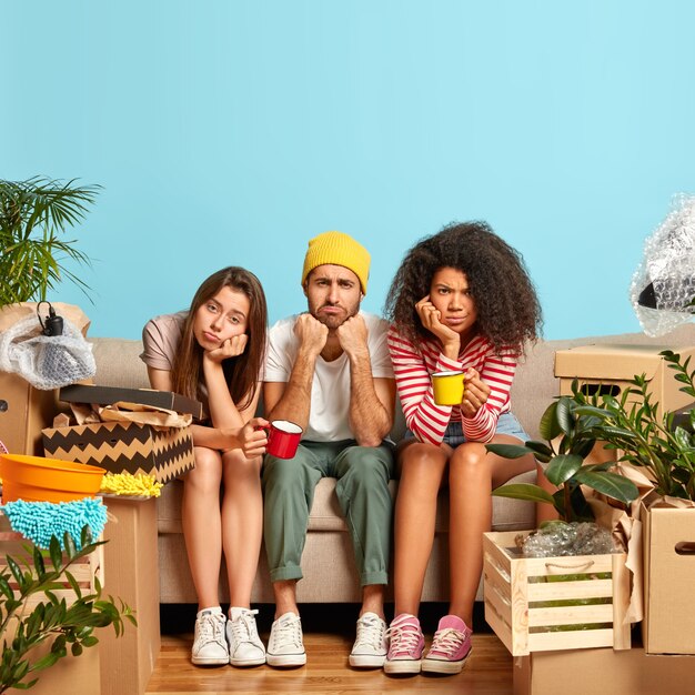 Vertical image of dissatisfied three multiethnic friends relocate in other place, have coffee break after unpacking belongings, look with dissatisfaction, sit on couch against blue wall in living room