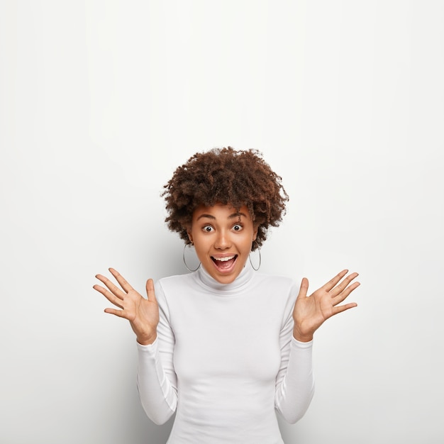 Vertical image of excited joyful curly haired woman spreads palms and looks happily , reacts on expensive present, stands alone against white wall, wears round earrings, poloneck