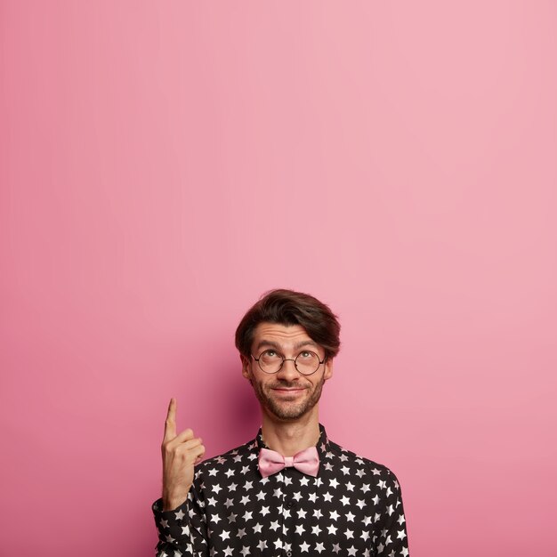 Vertical image of glad hipster with stubble points index finger above, dressed in fashionable elegant outfit