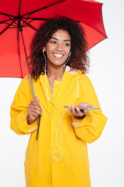 レインコートを着て傘の下に隠れて白で目をそらしながら彼女の電話で音楽を聴く幸せなアフリカ女性の垂直方向の画像