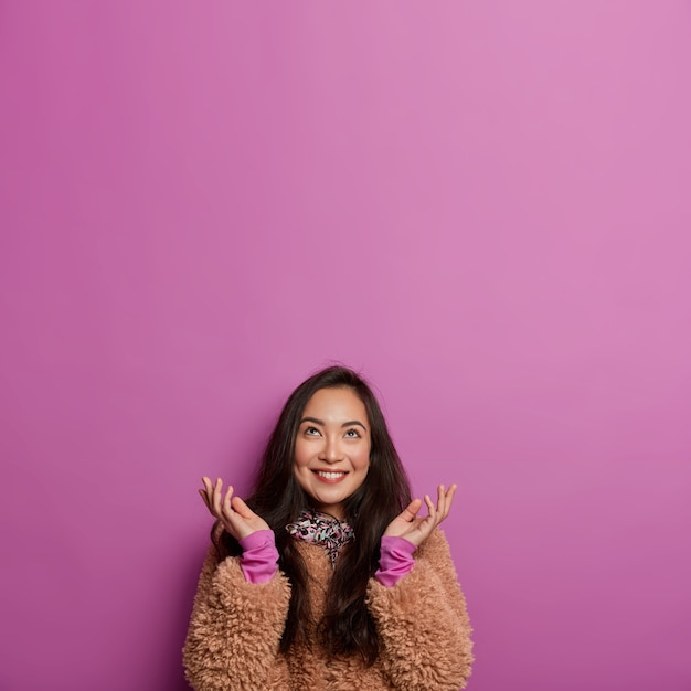 Vertical image of happy Asian lady looks hopefully above, spreads palms sideways, has tender smile, wears winter coat