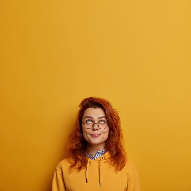 Vertical image of lovely ginger girl looks above, has pleased expression, raises head, enjoys sunny day, wears big glasses and sweatshirt