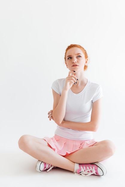 Vertical image of pensive ginger girl sitting on the floor and looking up