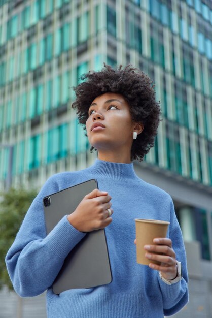 Вертикальное изображение задумчивой красивой женщины с вьющимися волосами, держащей цифровой планшет, а бумажный стаканчик с кофе в повседневной одежде позирует на открытом воздухе