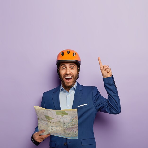 Free photo vertical image of satisfied male architect with blueprint, points above with index finger, has happy look, demonstrates something upwards, has idea in mind, wears protective hardhat, elegant suit