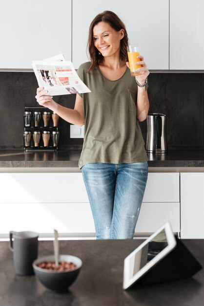 Vertical image of smiling casual woman reading newspaper and drinking juice on kitchen