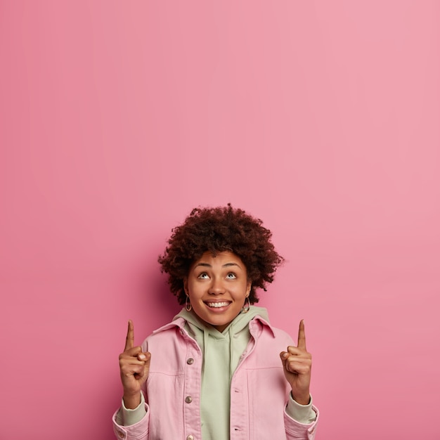 Vertical image of smiling optimistic curly woman points both index fingers above, has joyful expression, advertises cool promo upwards