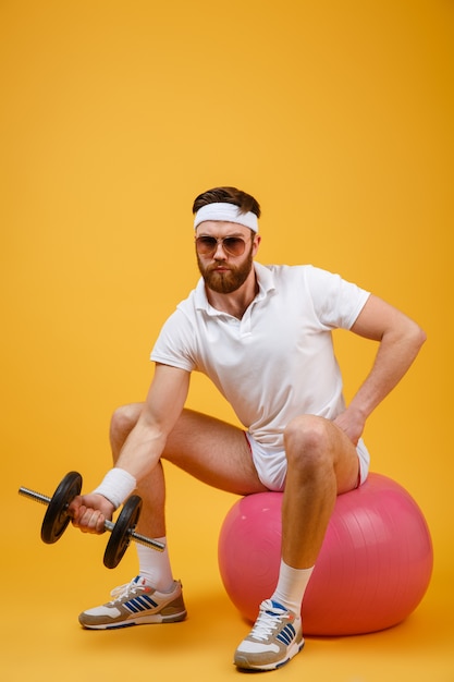 Vertical image of sportsman sitting on fitness ball with dumbbell