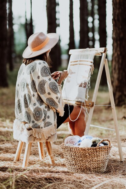 Vertical image of woman weaving a mat on a homemade loom with a basket of yarn