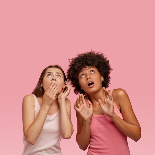 무료 사진 놀란 걱정스러운 두 여성의 수직 실내 촬영은 고개를 들고 위쪽으로 시선을 당겼으며 위층에서 공포증을 발견하고 텍스트를위한 여유 공간이있는 분홍색 벽 위에 서로 가까이 서 있습니다.