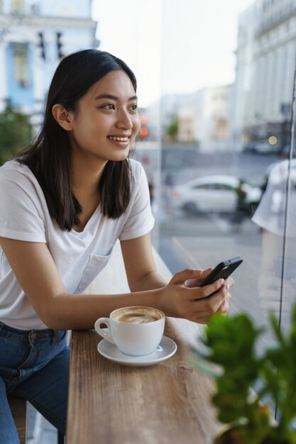 Vertical outdoor shot cheerful beautiful young modern asian girl in t-shirt, jeans, lean coffee table.