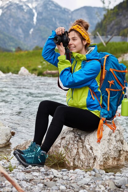 Vertical outdoor shot of cheerful female makes professional photos, sits on rocks near mountain river