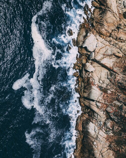 Vertical overhead shot of rocky shoreline next to a body of water with waves splashing the rocks