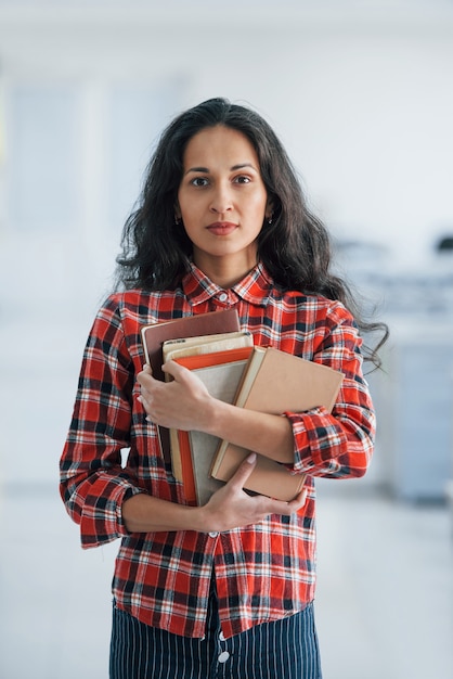 Вертикальное фото привлекательной молодой женщины, стоящей в офисе и держащей книги и документы