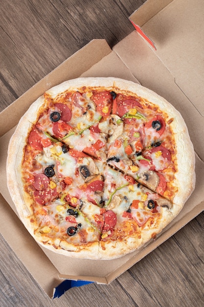 Vertical photo of fresh mixed pizza in pizza box over wooden table. 