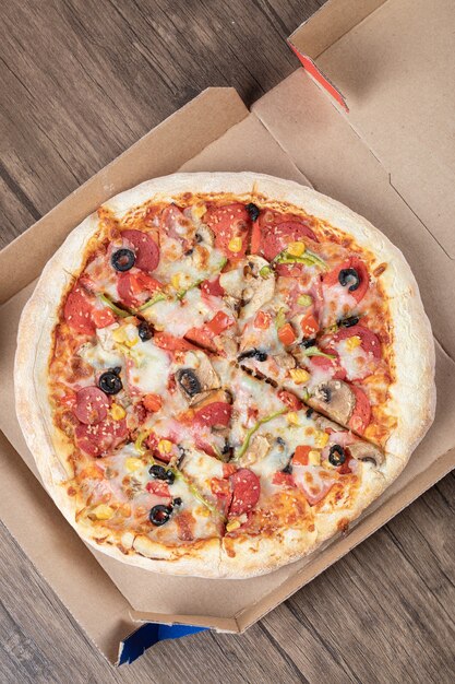 Vertical photo of fresh mixed pizza in pizza box over wooden table. 