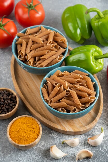 Vertical photo of raw penne pasta in bowls with fresh organic vegetables.