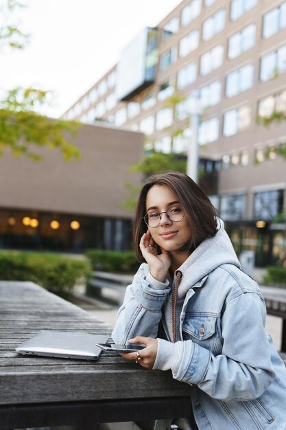 Вертикальный портрет очаровательной молодой студентки, сидящей в парке университетского городка возле места Wi-Fi, учащегося в ожидании друга, использующего ноутбук и мобильный телефон, улыбающегося веселой камеры