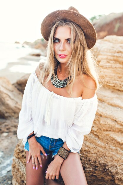 Vertical portrait of attractive blonde girl with long hair sitting on stone  on deserted beach.  She is looking to the camera.