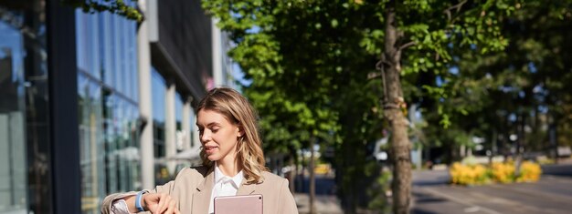 Free photo vertical portrait of beautiful businesswoman in suit holds her digital tablet looks at watch on