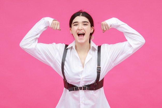 Vertical portrait of beautiful motivated brunette squeezing her biceps and stands on pink background