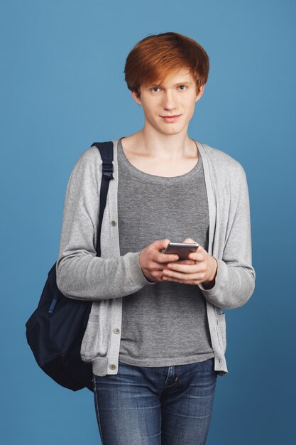 Vertical portrait of beautiful serious young student guy with red hair in casual outfit with black backpack chatting with girlfriend by phone,  with calm expression