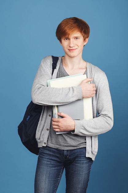 Vertical portrait of beautiful young cheerful male student in casual outfit with backpack smiling, holding lot of notebooks in hands,  with satisfied expression.