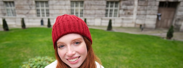 Free photo vertical portrait of cute redhead female student in red hat and warm gloves sits in park on bench