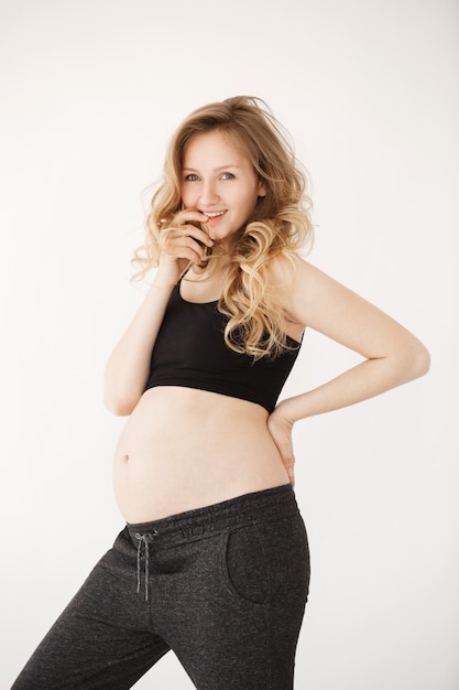 Vertical portrait of good-looking charming young mother with light wavy hair in sport clothes smiling, holding hand near lips, looking at her husband with flirty face expression. Pregnancy and materin