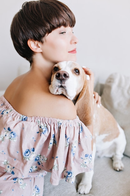 Vertical portrait of graceful lightly tanned woman gently holding her beagle dog on white wall