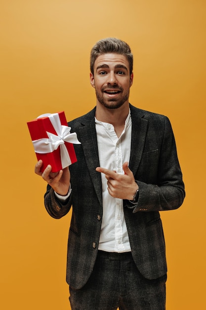 Vertical portrait of handsome bearded man in checkered jacket pants and white shirt points at red gift box on orange background