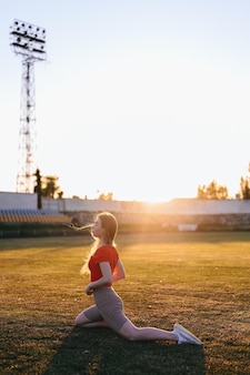 Вертикальный портрет девушки в спортивной одежде, делающей растяжку на солнце на закате и смотрящей в камеру