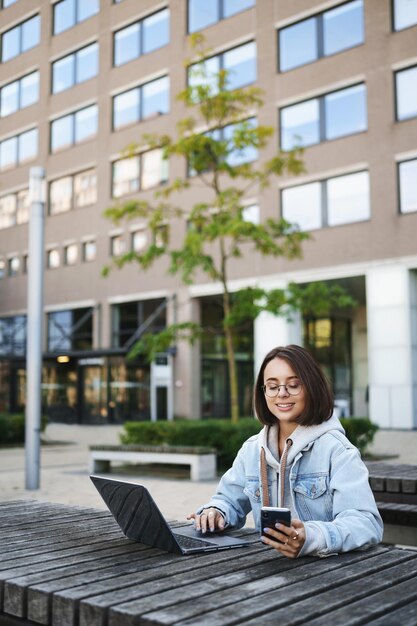 Vertical portrait of smiling cute young woman sitting oudoors surrounded by buildings using mobile phone and laptop smiling at smartphone display working on remote freelancing