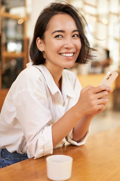 Vertical portrait of stylish asian woman sitting in cafe drinking coffee and using smartphone