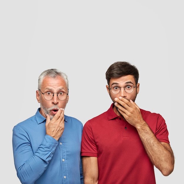 Free photo vertical portrait of two men colleagues being stupefied to find out about raising price, cover mouthes, stand closely, isolated over white wall. emotional handsome father and son of different age