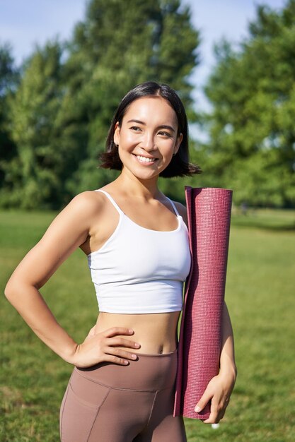 Vertical portrait of young asian fitness girl walks with rubber mat for yoga goes on training sessio