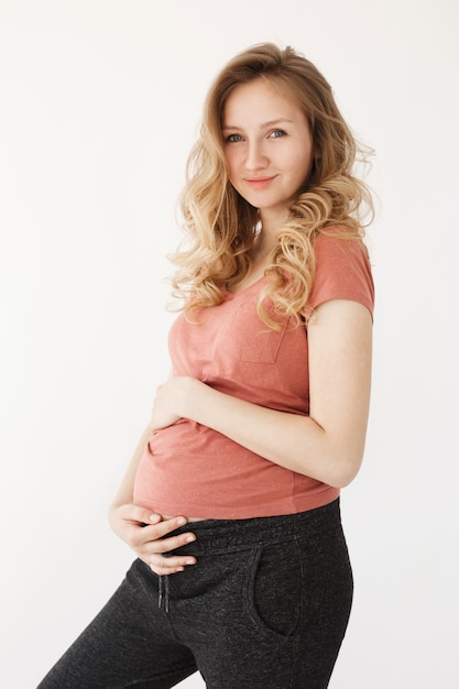 Vertical portrait of young good-looking cheerful european mother with long blonde hair in casual clothes holding pregnant belly with hands