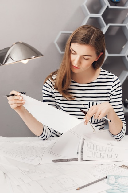 Free photo vertical portrait of young good-looking female designer with brown hair in striped shirt, looking at papers with serious expression, working on new clothes designs for fashion show.